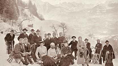 Historische Rodelgesellschaft in Schruns im Montafon mit Schlitten, Wintersportlern und verschneiter Berglandschaft.