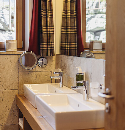 Waschbereich der Familiensuite Edelweiß im Genusshotel Vermala mit Aufsatzwaschbecken, Kosmetikspiegel und Fenster.