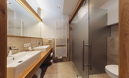 Badbereich der Suite Brunella im Genusshotel Vermala mit Doppelwaschbecken, Dusche, WC und moderner Natursteinoptik.